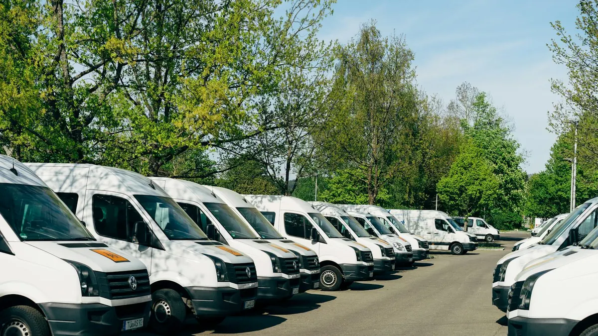 A row of white service vans parked nose-out along a yard, trees and sky behind them.