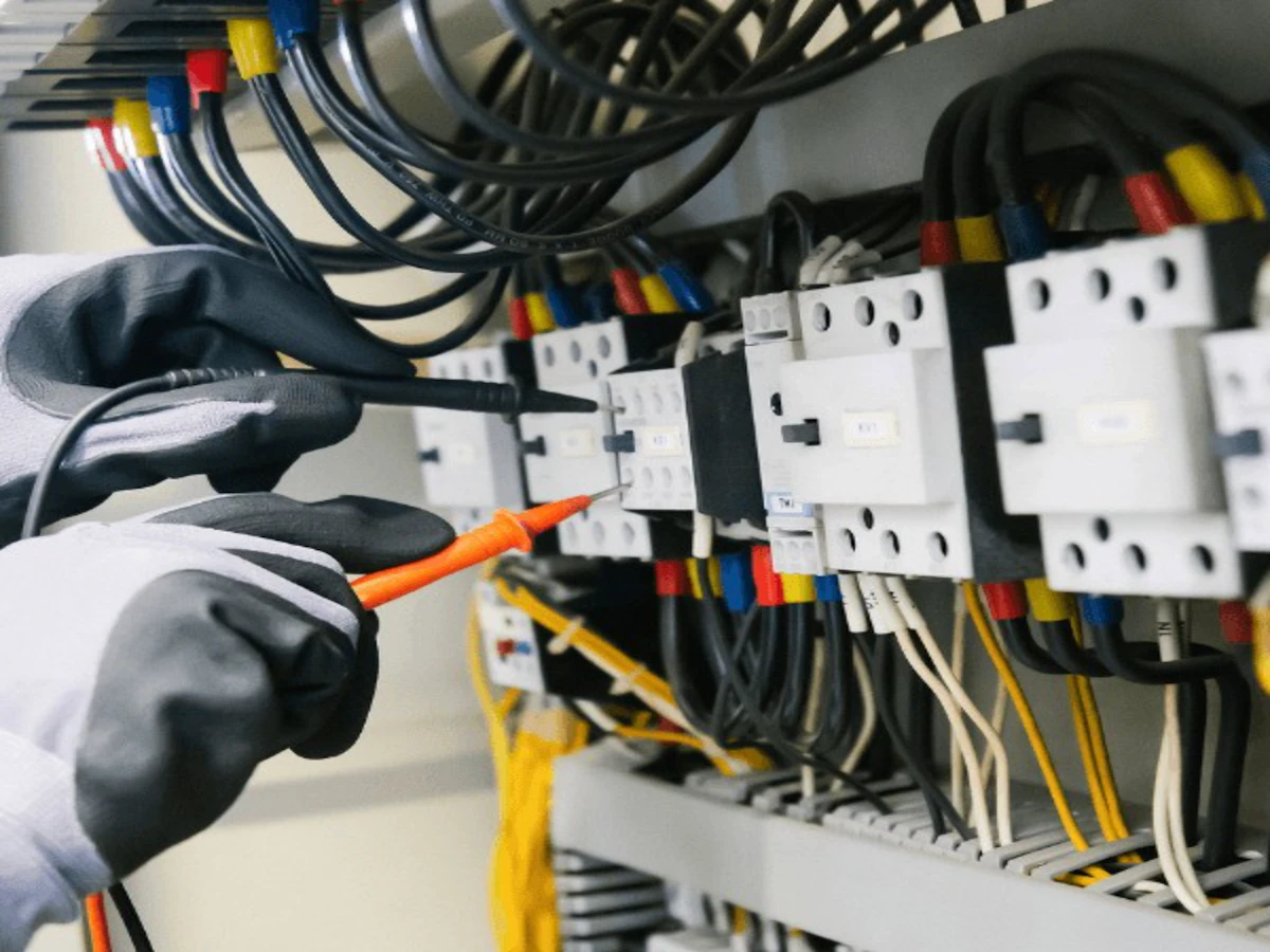 One pair of gloved hands holding multimeter probes against a row of contactors in an open control panel.