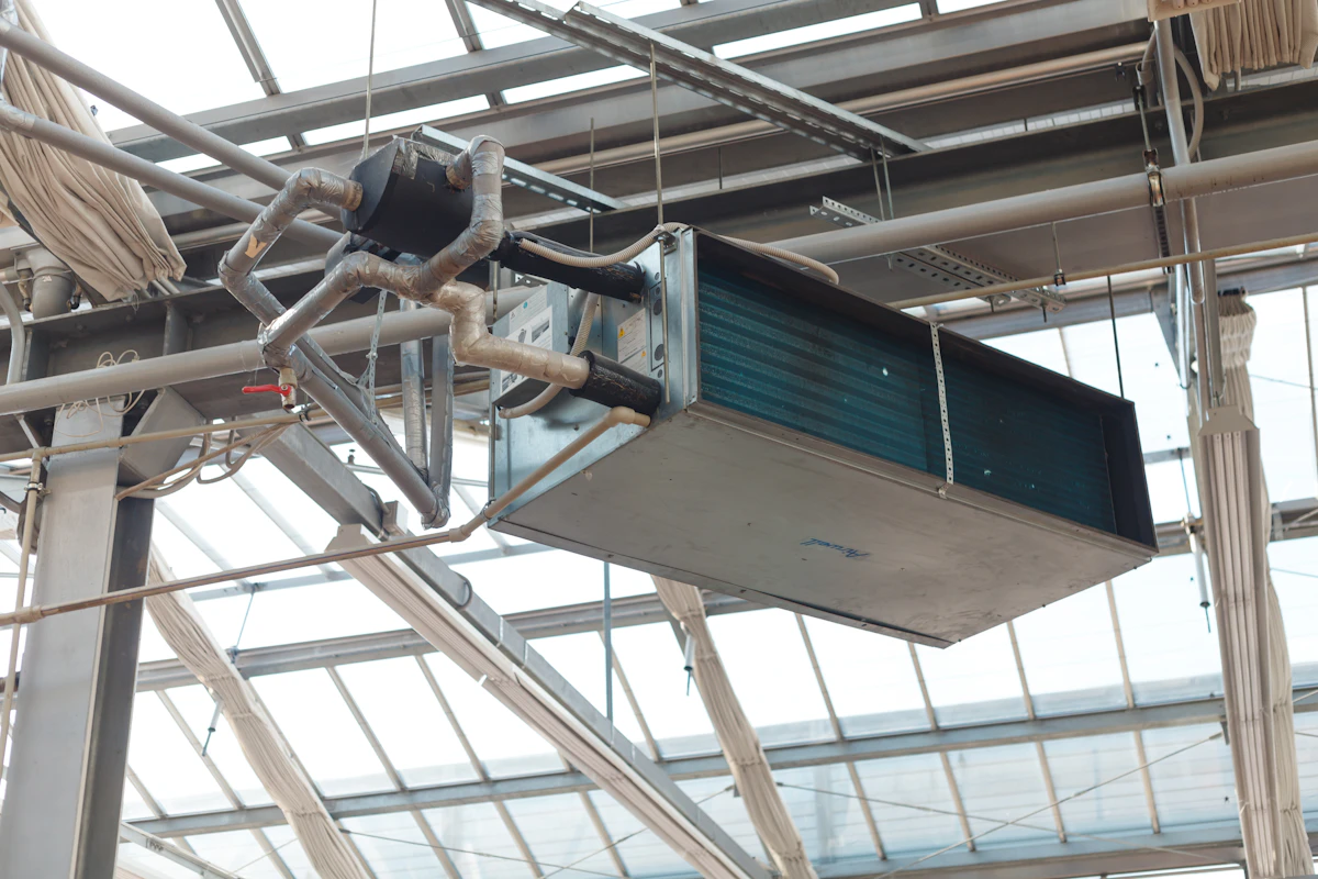 A ceiling-suspended air handler and its insulated refrigerant lines, hung from the steel roof structure of a glass-roofed building.