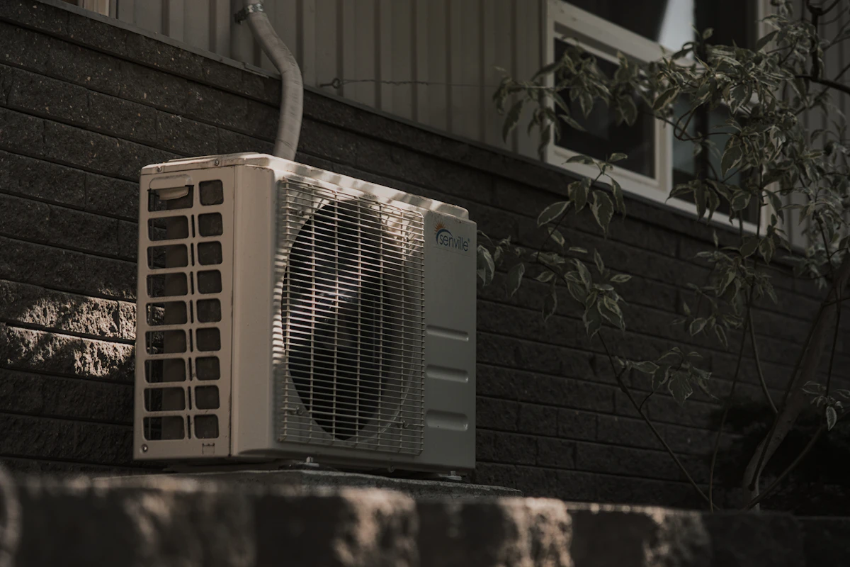 A residential outdoor condenser unit sitting on a stone ledge against the dark brick wall of a house, its refrigerant lines running up beside it.