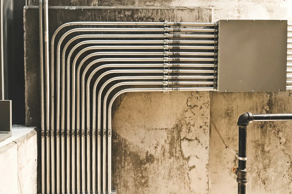 Ten stainless pipe runs turning together into a panel on a concrete wall.