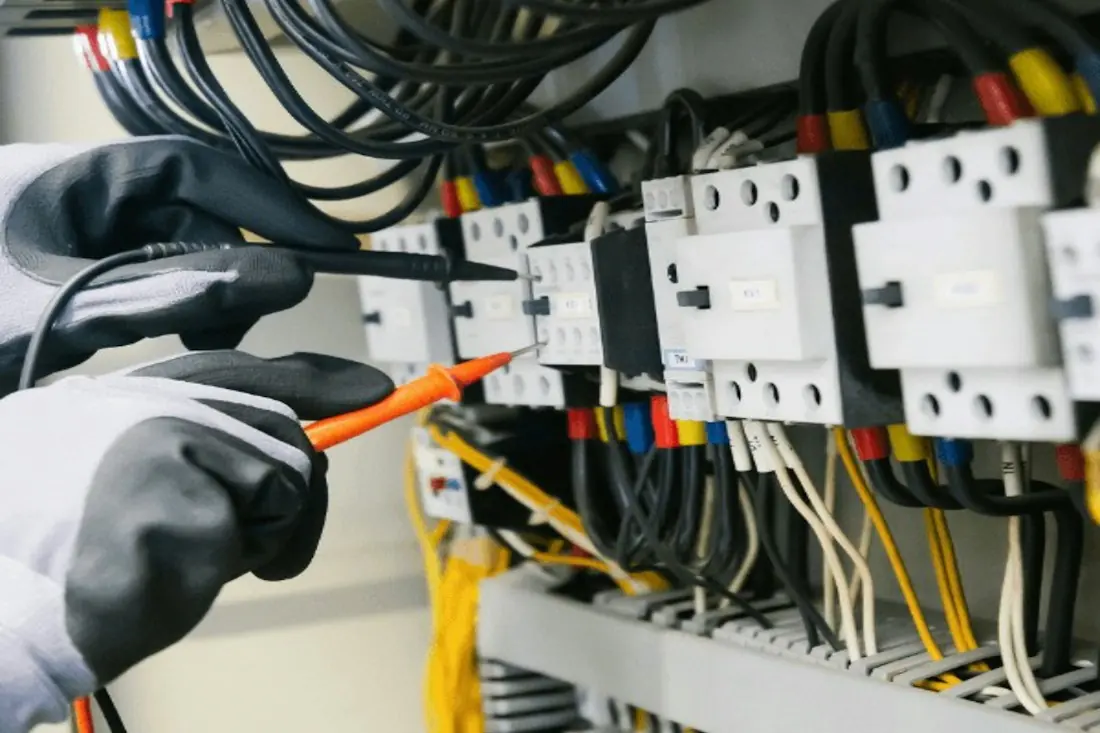 A gloved hand holding an orange test probe against one terminal in a row of breakers and coloured conductors.