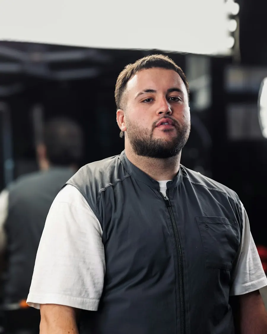A bearded electrician in a work vest over a white t-shirt, looking straight at the camera, a workshop out of focus behind him.