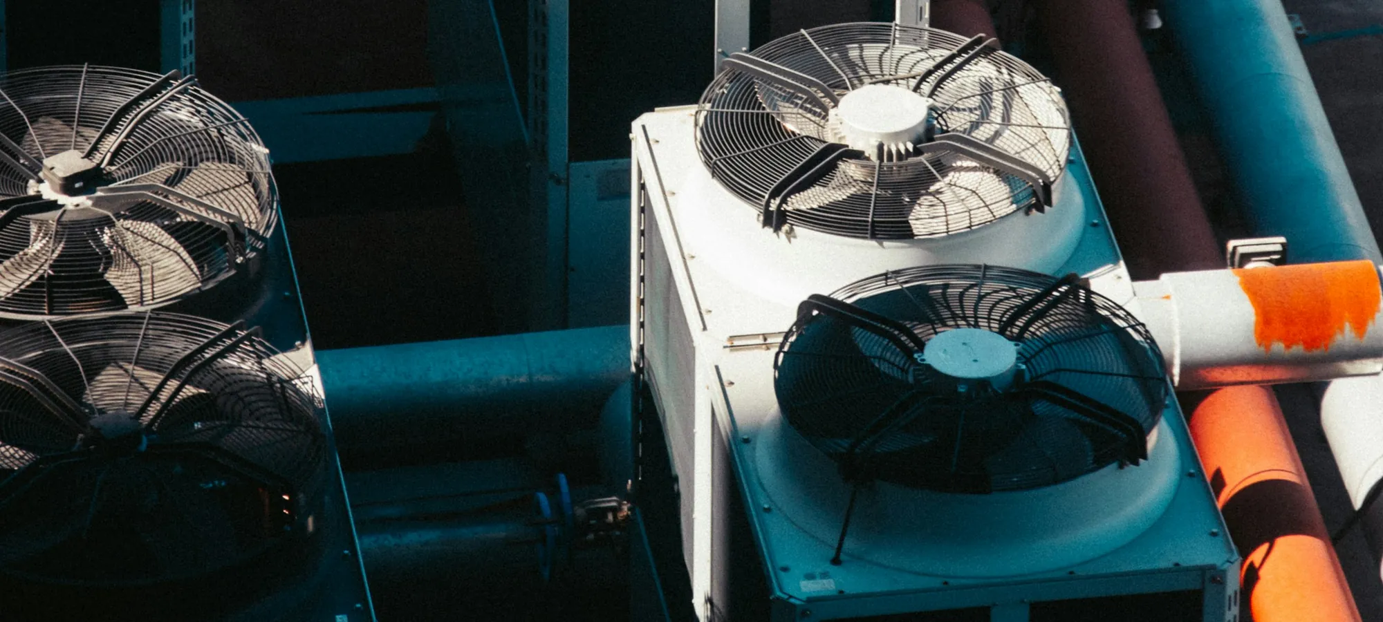 Looking down on a rooftop plant: three large condenser cabinets with fan grilles, insulated pipe runs crossing between them.