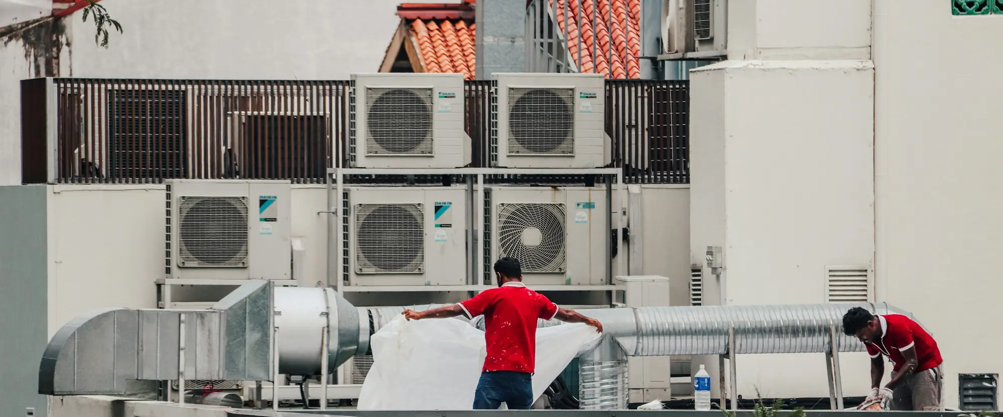 Two technicians on a flat roof fitting insulation along a run of galvanised duct, beside a bank of eight condensing units.