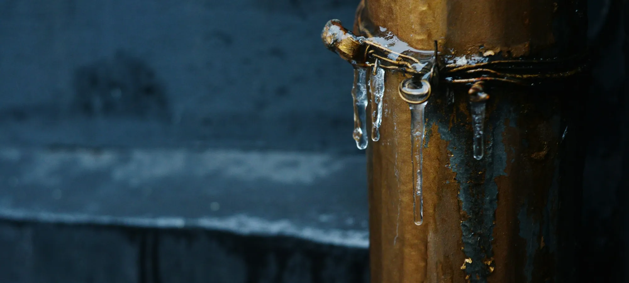Icicles hanging from the wire strap on a brass downpipe against a dark slate wall.