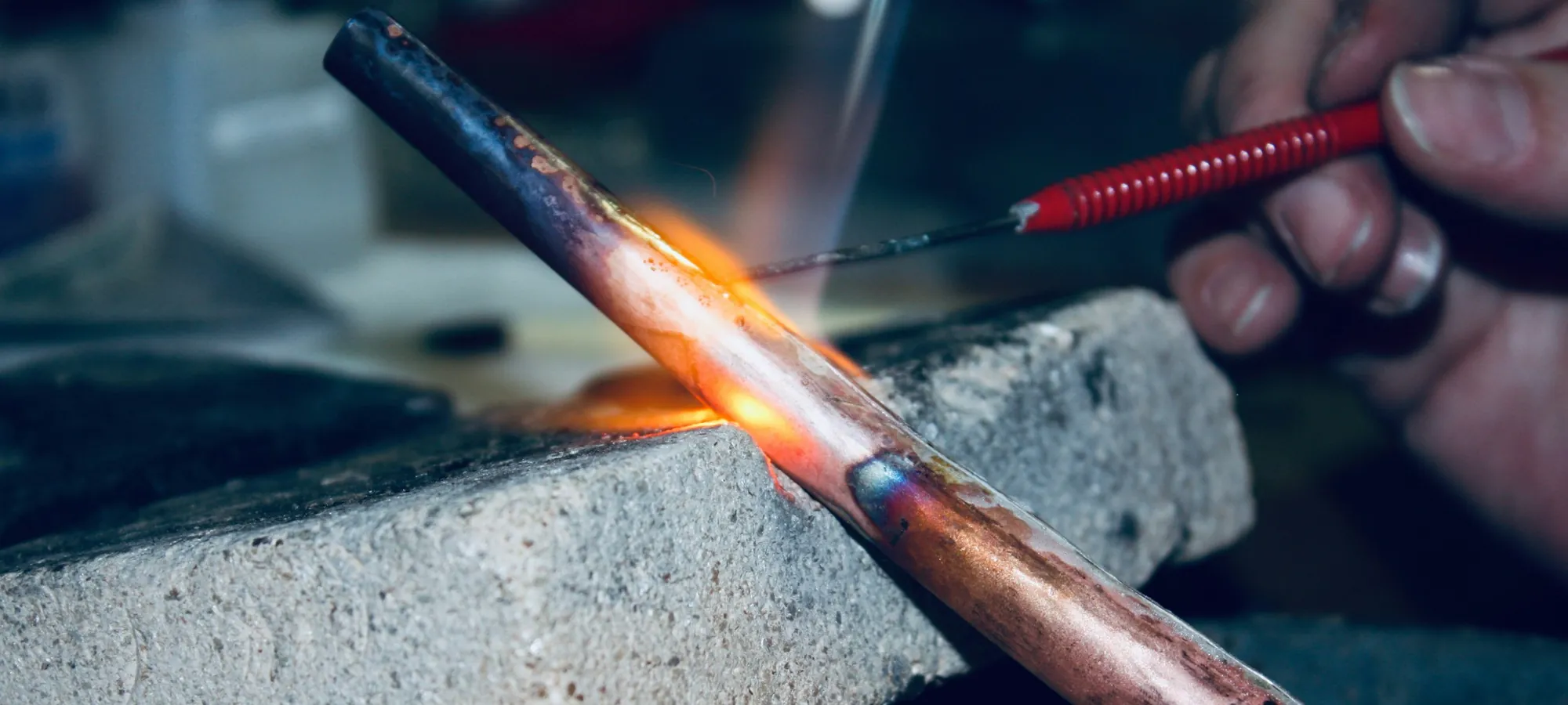 A blue torch flame and filler rod on a copper pipe joint resting on a fire block.